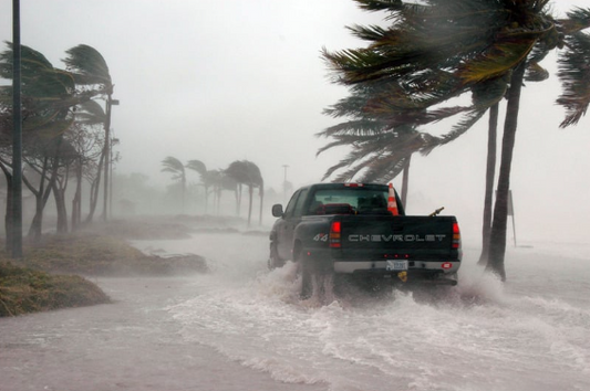 Truck driving down a road through flooding whilst the trees are blowing in a hurricane.