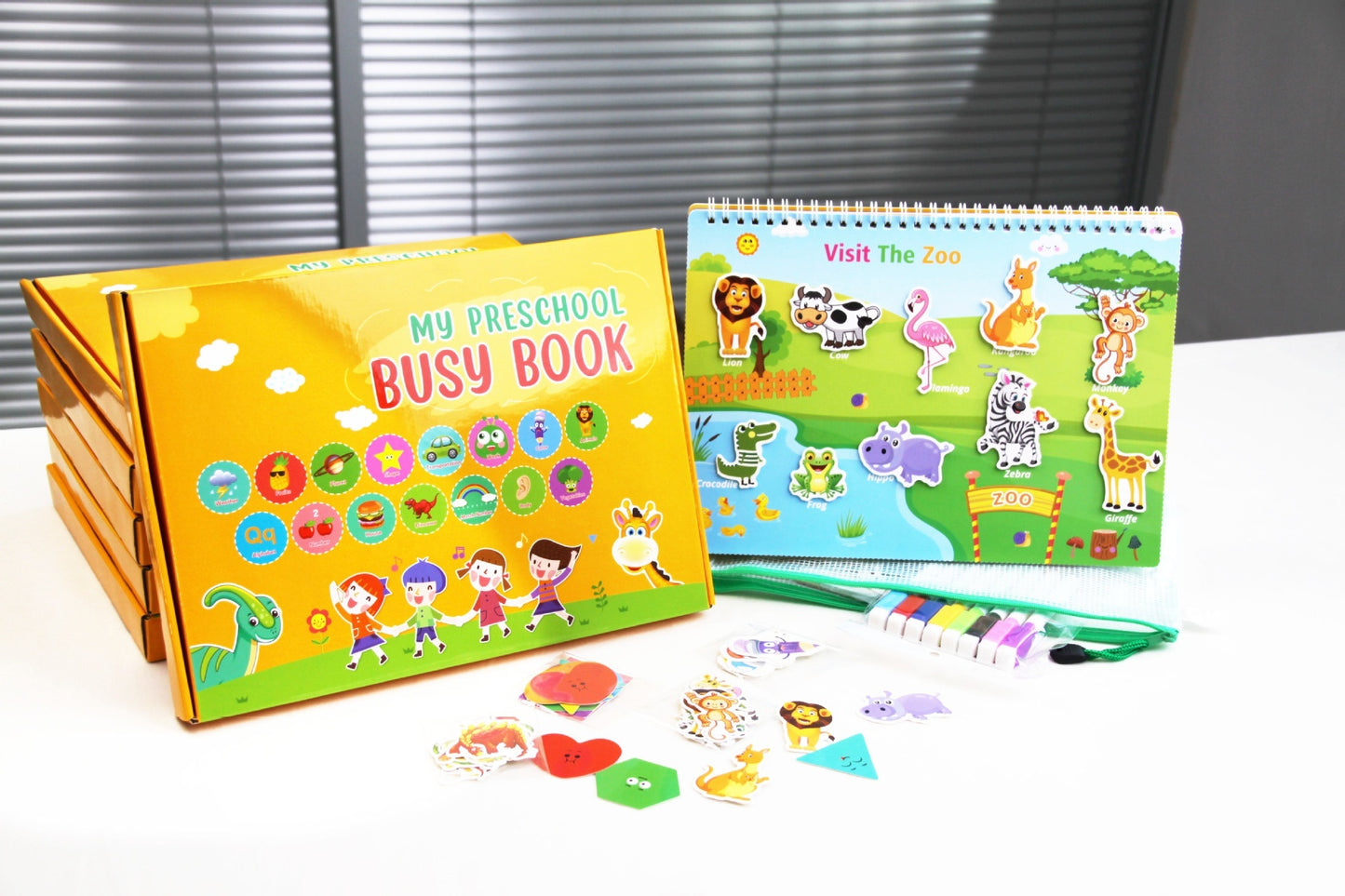 This shows the front of the box and it says My Preschool Busy Book. the book is on the visit the zoo page. the book is stood up on the table and next to it is animal cards, coloured pens the storage box and bag.
