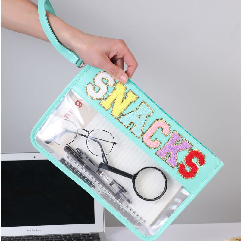 A grey wall and computer desk in the background and in the foreground a hand is holding up a clear and transparent light blue lined pouch bag with the word snacks across the top in colourful letters that are lined with glittery gold. In the bag you can see multiple different items. Also there is a wrist strap attached to the zipper of the snack bag that matches the light blue lining of the bag.