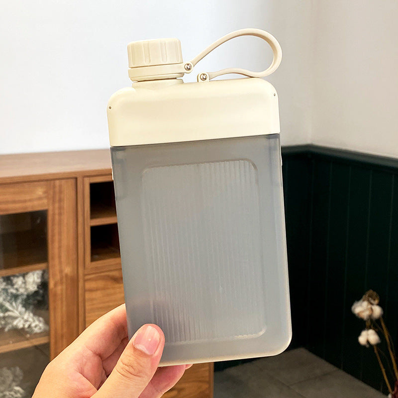 Clear flat square water bottle with beige cap held in a hand against a neutral background