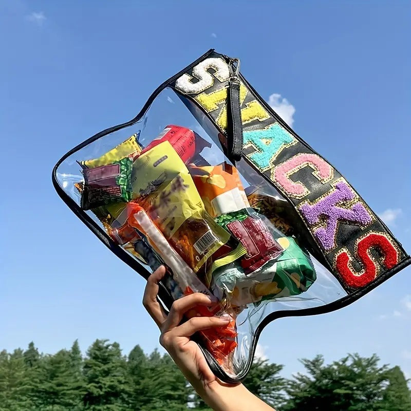 A blue sky and green trees background and in the foreground a hand is holding up a clear and transparent Black lined pouch bag with the word snacks across the top in colourful letters that are lined with glittery gold. In the bag you can see multiple different snacks. Also there is a wrist strap attached to the zipper of the snack bag matches the black lining of the bag.