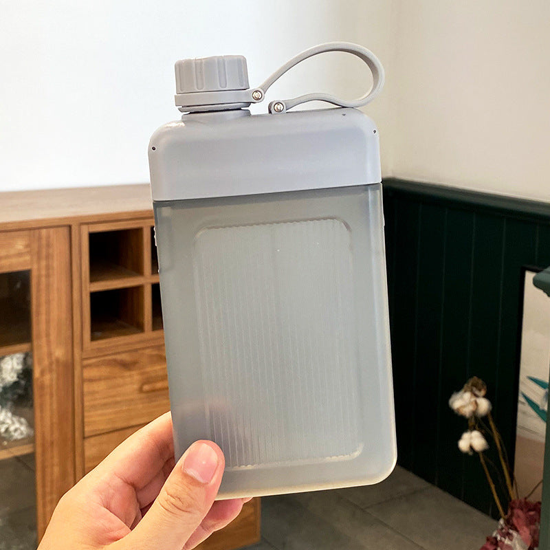 Person holding a translucent flat square water bottle with a grey cap indoors.