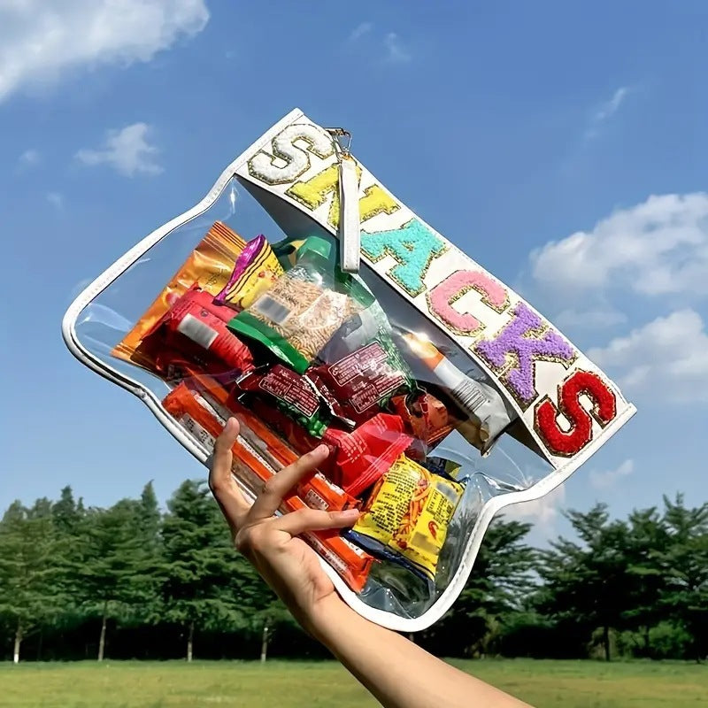 A blue sky and green trees background and in the foreground a hand is holding up a clear and transparent white lined pouch bag with the word snacks across the top in colourful letters that are lined with glittery gold. In the bag you can see multiple different snacks. Also there is a wrist strap attached to the zipper of the snack bag that matches the white lining of the bag.