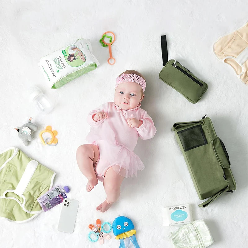 shows a baby surrounded by baby stuff that could go into the bag, nappies, toys, wipes, phone, tether, rattle, baby clothes.