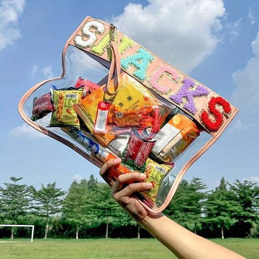 A blue sky and green trees background and in the foreground a hand is holding up a clear and transparent  
Pink lined pouch bag with the word snacks across the top in colourful letters that are lined with glittery gold. In the bag you can see multiple different snacks. Also there is a wrist strap attached to the zipper of the snack bag that matches the pink lining of the bag.
