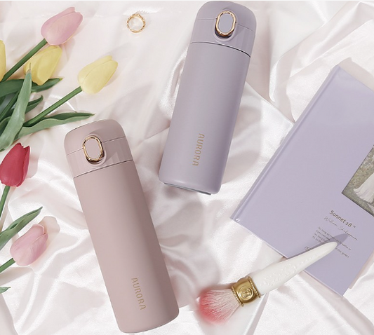 The Aurora Stainless Steel Insulated Bottle in this picture we have 2 of the bottles in Light Su Pink and Hyacinth Purple laying on white silk sheets with flowers, make up brush and book.