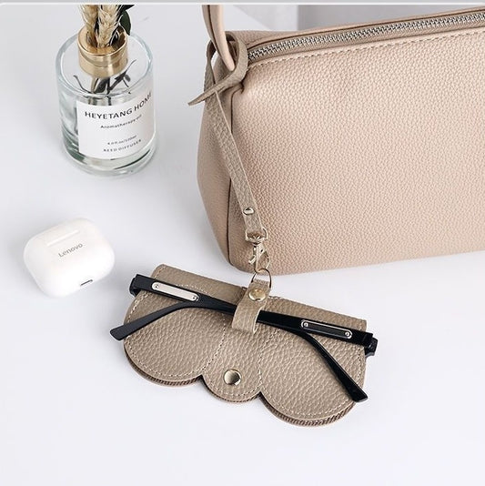 A beige handbag on a white counter showing how the clip on glasses holder is used with a pair of glasses in the holder and the holder attached to the bag. A great travel glasses case.